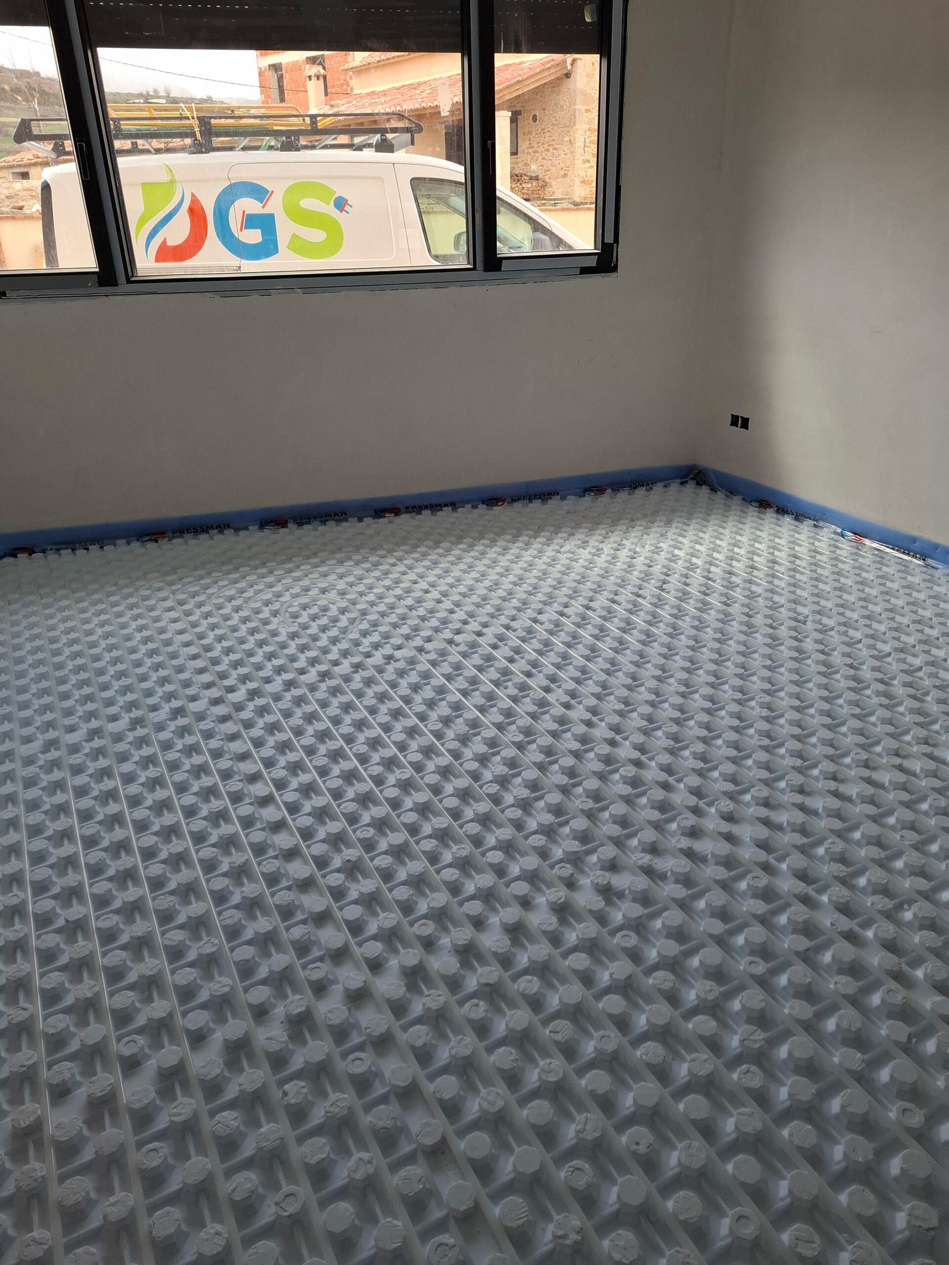 Habitación en obra con suelo radiante instalado y furgoneta de la empresa DGS visible por la ventana.